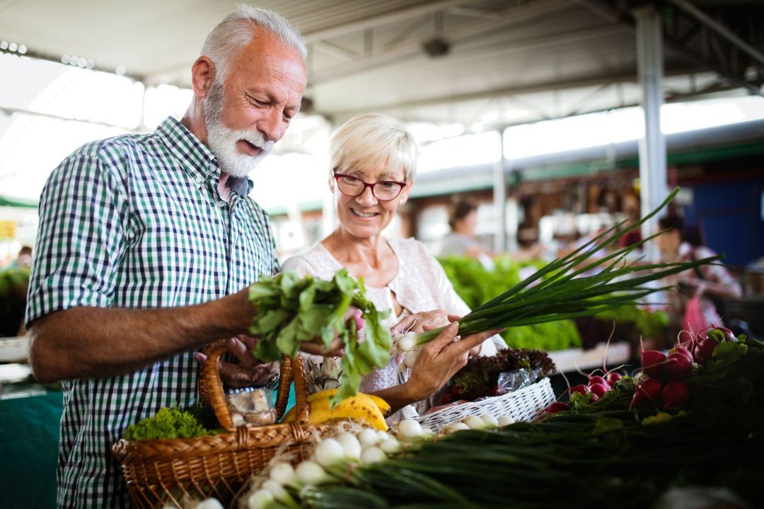 Meridian at Wylie Older couple smiling and shopping for fresh vegetables at an outdoor farmer’s market.