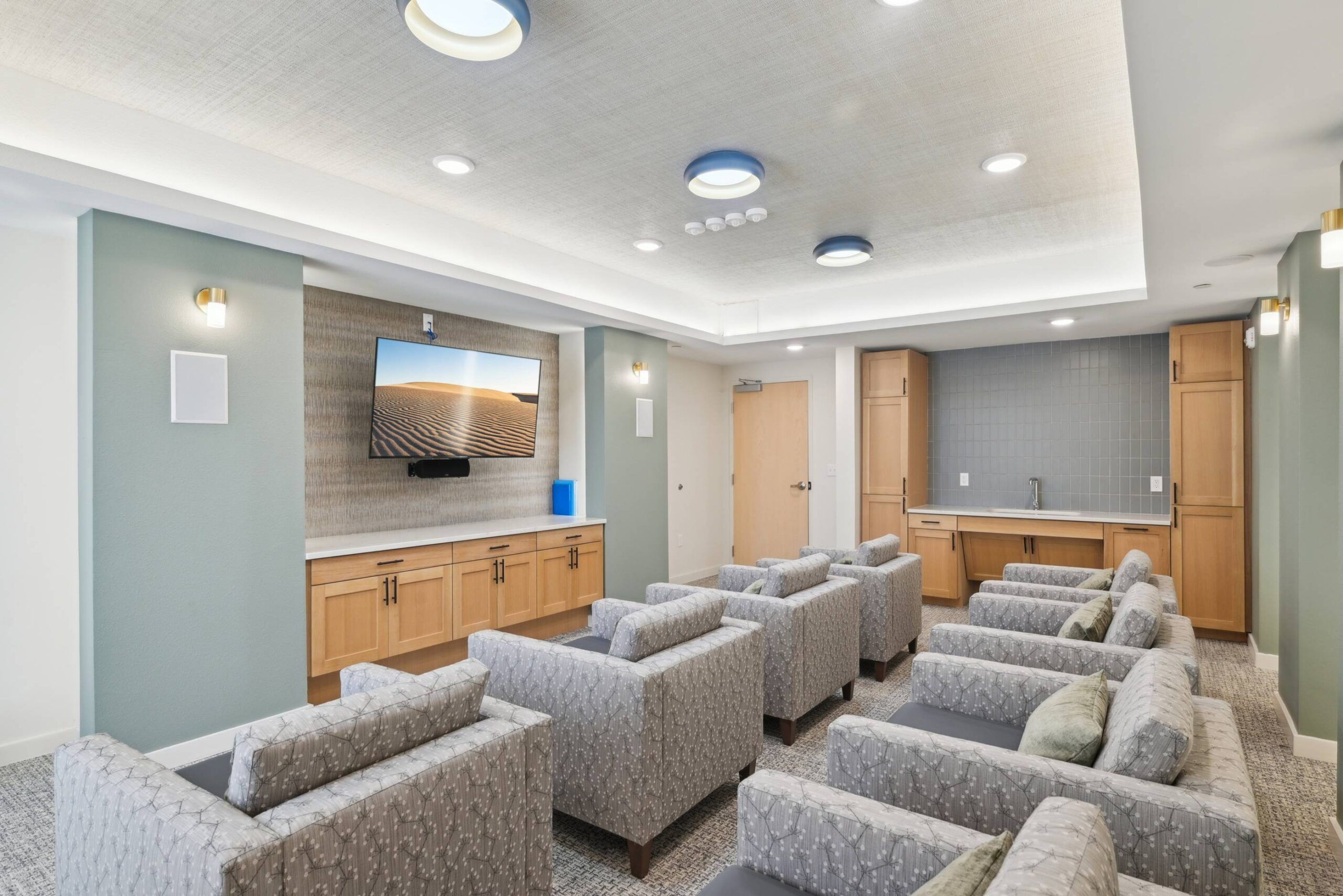 Meridian at Wylie Modern media room with patterned armchairs facing a wall-mounted TV, wood cabinets, and a small kitchenette.