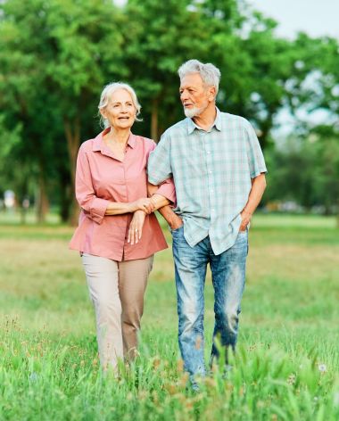 Meridian at Wylie An elderly couple walks arm in arm through a grassy park, smiling on a sunny day.