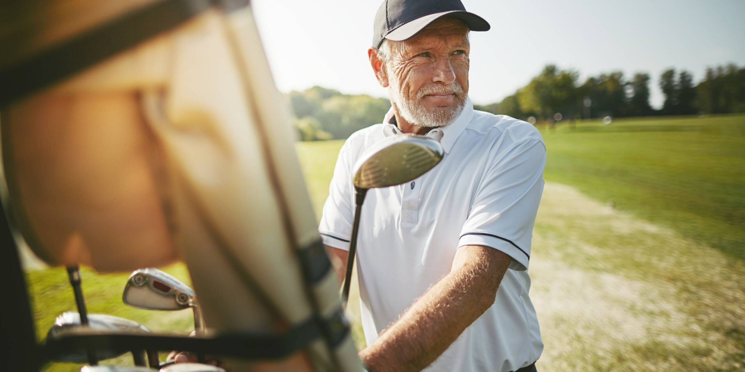 Meridian at Wylie Older man in a white polo and cap stands by a golf cart on a sunny golf course, holding a golf club.