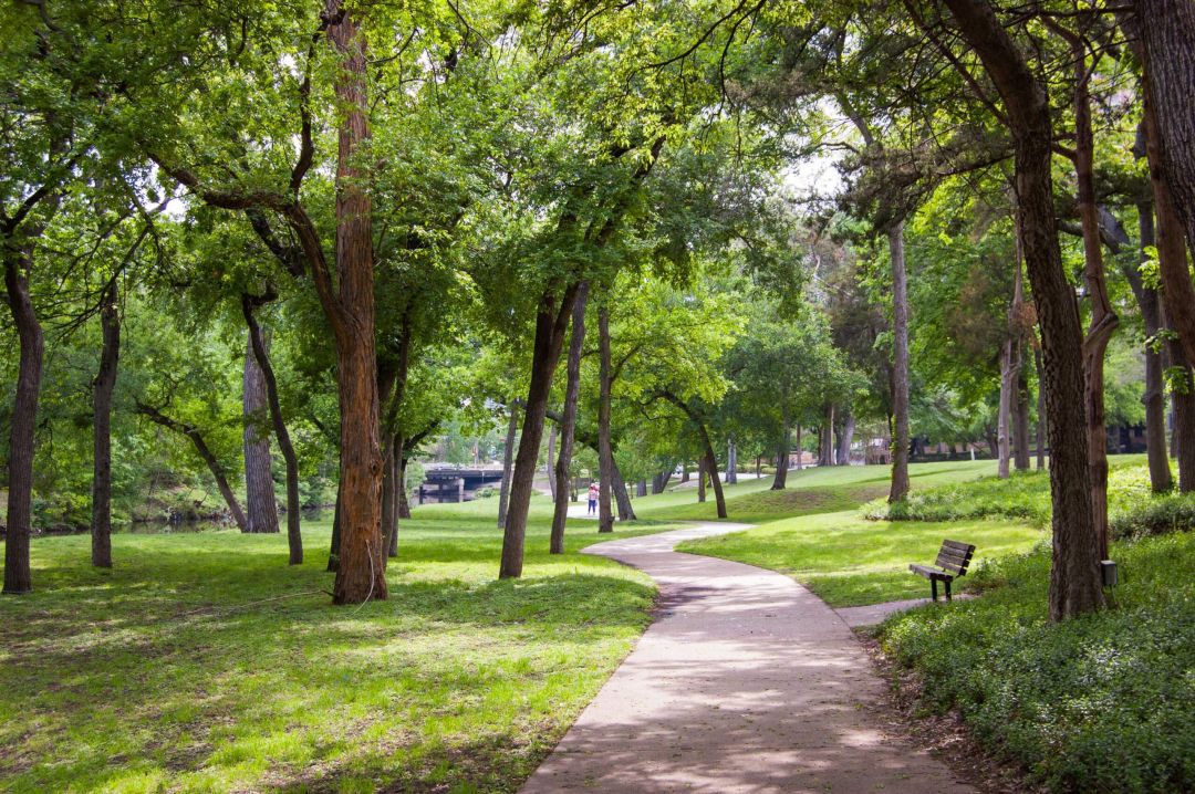 Meridian at Wylie A winding path through a green park with trees, grass, and a wooden bench on a sunny day.